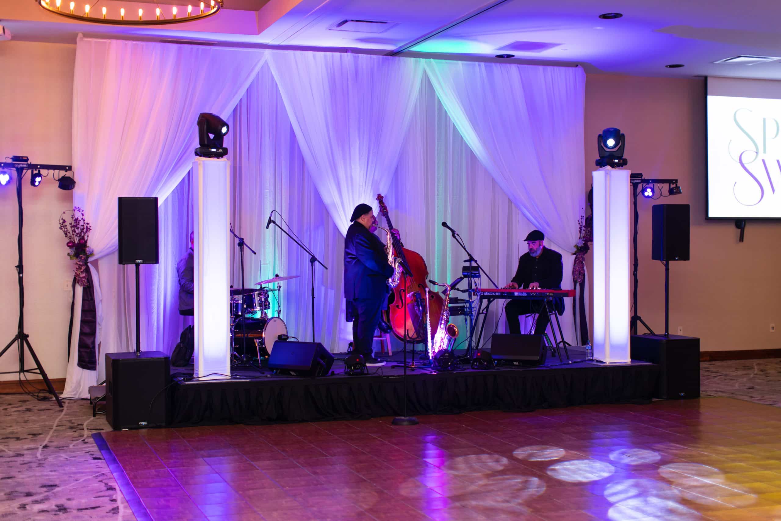 Live band on a draped gala stage with colored lighting, illuminated towers, speakers, and projection screens.