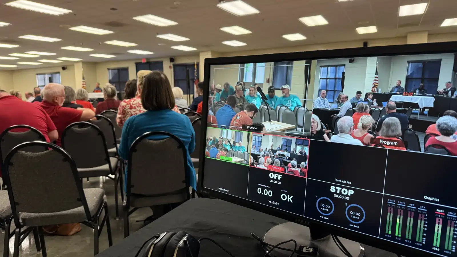 Video production monitor showing two camera feeds during a public meeting with an audience